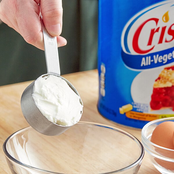 A person using a measuring cup to scoop Crisco shortening into a clear bowl.