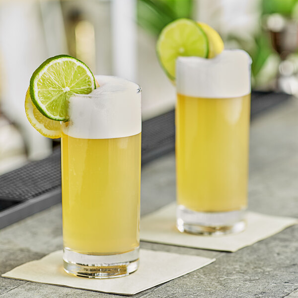 Two glasses of yellow liquid with lime slices on top in front of a yellow DaVinci Gourmet cocktail mixer.