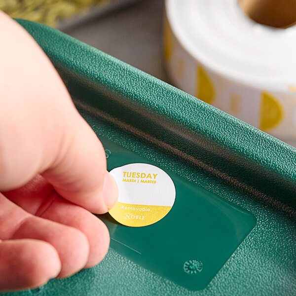 A hand holding a roll of Noble Products Tuesday Day of the Week stickers on a green tray.