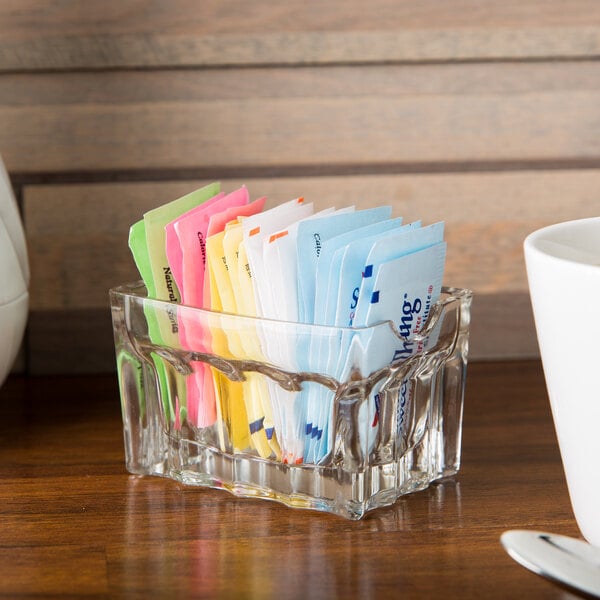 A Tablecraft fluted glass sugar caddy filled with colorful sugar packets on a table.