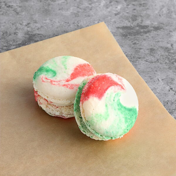 Two Macaron Centrale cranberry pistachio macarons with green and red icing on a piece of paper.