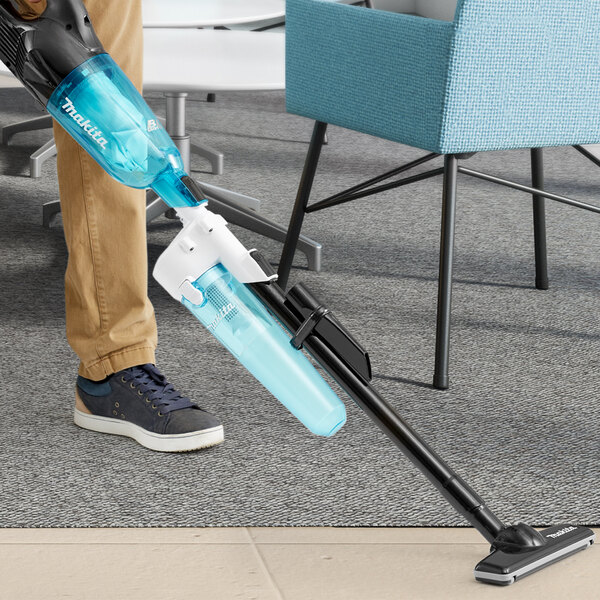 A person using a Makita white cyclonic stick vacuum to clean a blue chair