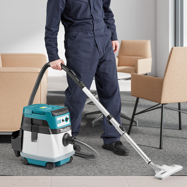 A person in blue overalls using a Makita XCV23Z vacuum cleaner with a hose to vacuum a carpet.