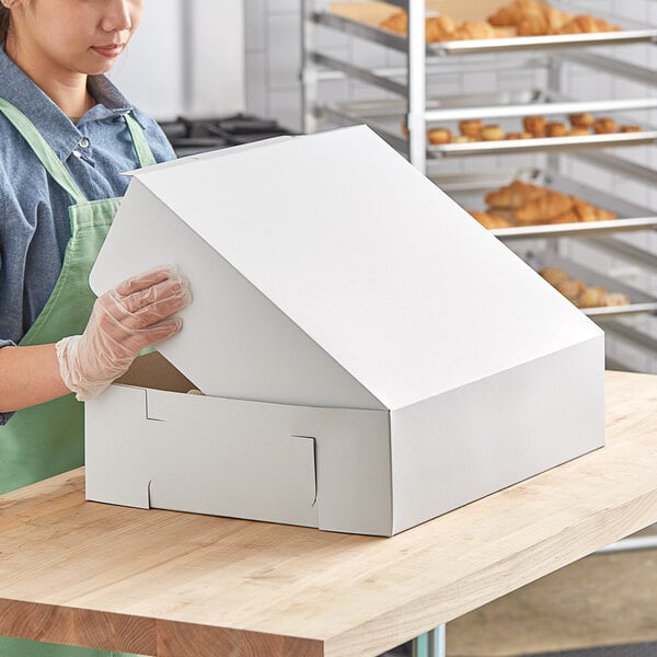 A woman wearing gloves and an apron opening a white Baker's Mark half sheet cake box.