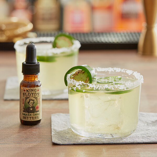 A bottle of King Floyd's Green Chili Bitters on a table next to two glasses of margaritas.