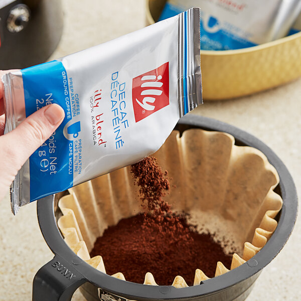 A hand pouring illy Decaf Classico coffee powder from a blue bag into a coffee filter.
