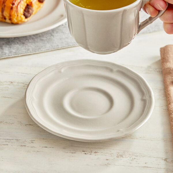A hand holding an Acopa Condesa warm gray porcelain saucer with a cup of tea over a plate of food.