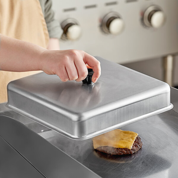A person using an American Metalcraft aluminum square basting cover to cook a hamburger on a metal surface.