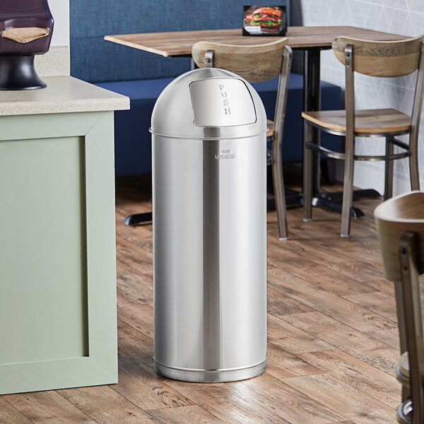 A Lancaster Table & Seating stainless steel decorative trash can with a push door lid on a wood floor.