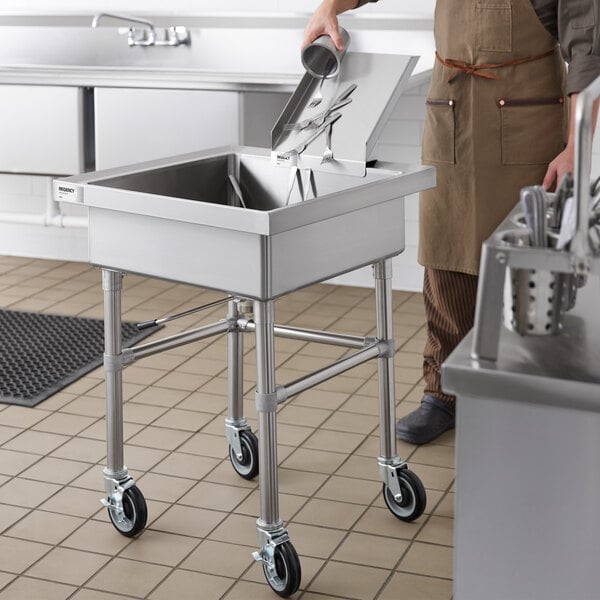 A stainless steel mobile soaking sink with a detachable chute, shown in a commercial kitchen setting with utensils being placed inside.