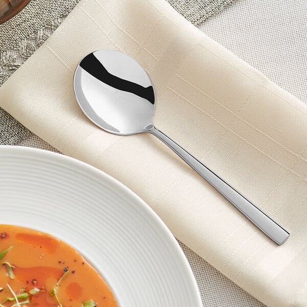 An Acopa stainless steel bouillon spoon on a napkin next to a bowl of soup.