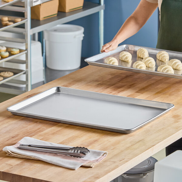 A large, empty aluminum sheet pan sits on a wooden countertop, with another pan holding dough pieces being carried by a person in the background.