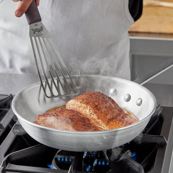 A hand using a spatula to cook food in a Choice aluminum fry pan on a stove.