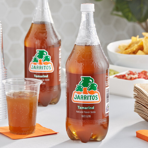 A close up of a bottle of Jarritos Tamarind soda with brown liquid next to a clear plastic cup of brown liquid.