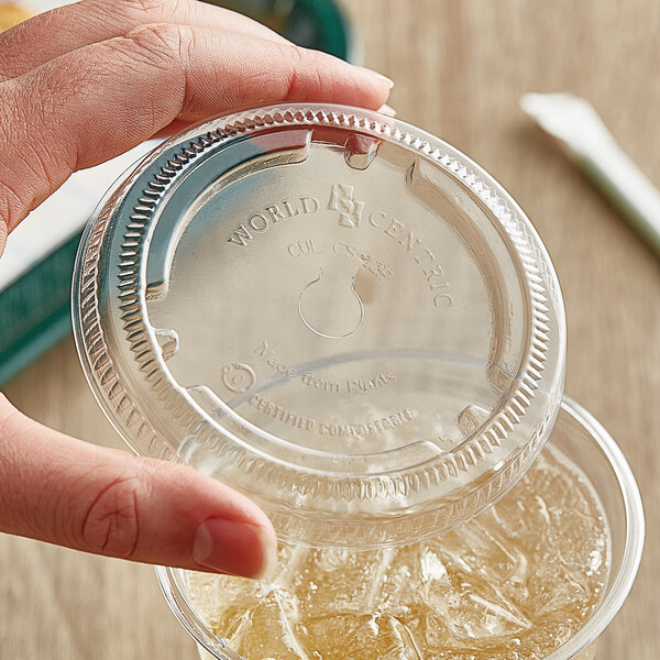 A hand holding a World Centric clear plastic lid with a straw slot on a plastic cup.