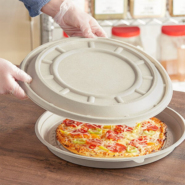 A gloved hand placing a World Centric compostable pizza lid on a container of pizza.