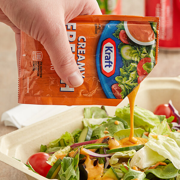 A hand pouring a Kraft Creamy French Dressing packet over a salad on a table.