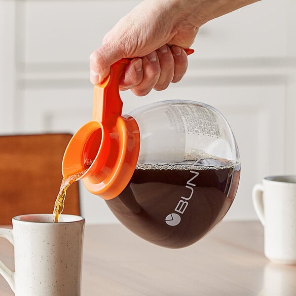 A hand with an orange handle pouring coffee into a white cup.