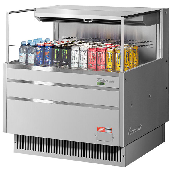 A Turbo Air stainless steel horizontal refrigerated curtain merchandiser on a counter filled with cans of soda.