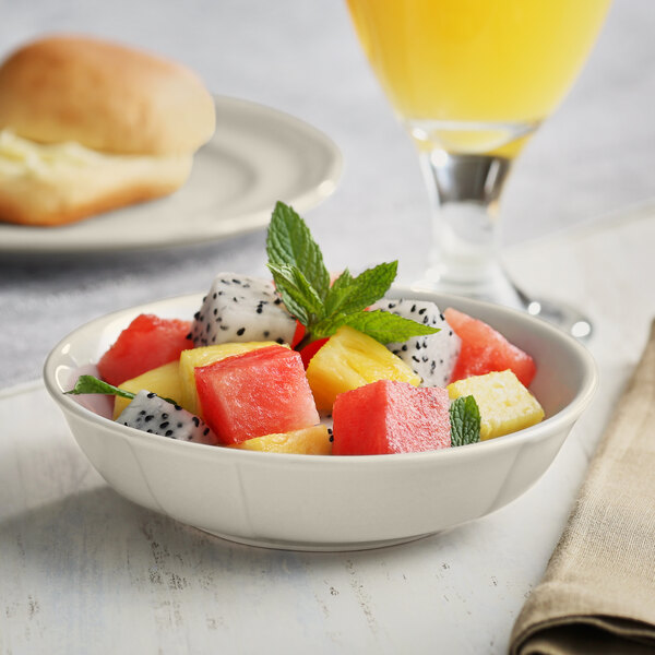 An Acopa Condesa scalloped porcelain fruit dish filled with fruit salad on a table next to a glass of juice.