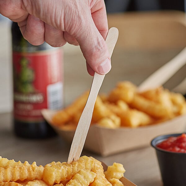 A hand holding a Royal Paper Eco-Friendly Wood French Fry Fork over french fries.