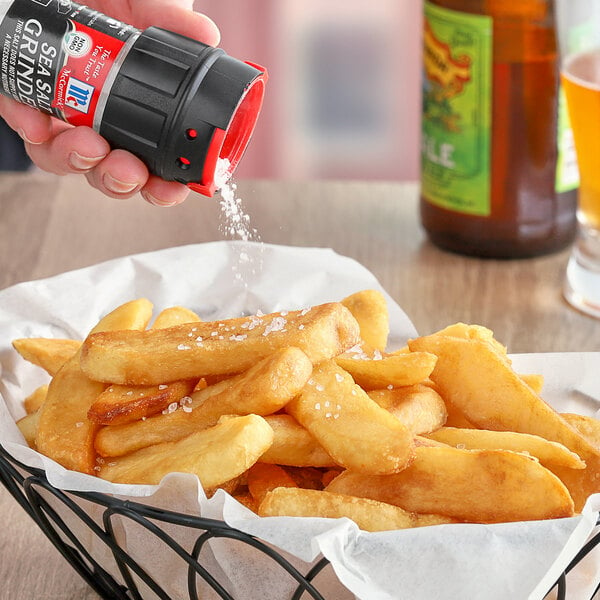 A hand using a black and red McCormick Sea Salt grinder to sprinkle salt on a basket of french fries.