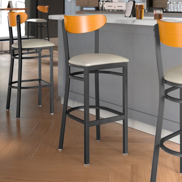 Lancaster Table & Seating Boomerang Series bar stools with light gray vinyl seats and cherry wood backs next to a restaurant counter.