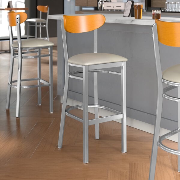 A group of Lancaster Table & Seating Boomerang Series bar stools with light gray vinyl seats and cherry wood backs.