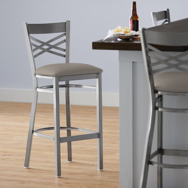 A Lancaster Table & Seating clear coat finish cross back bar stool with a dark gray vinyl padded seat.