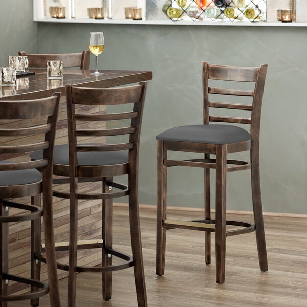 A Lancaster Table & Seating wood ladder back bar stool with a dark gray vinyl seat.