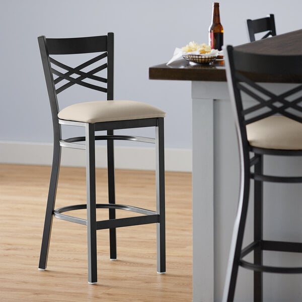 A Lancaster Table & Seating black cross back bar stool with a light gray cushion on the seat.