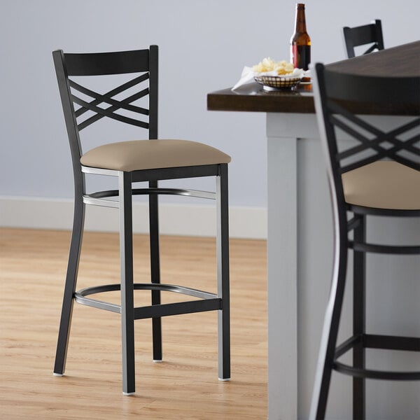 A Lancaster Table & Seating black cross back bar stool with taupe vinyl padded seat next to a table.
