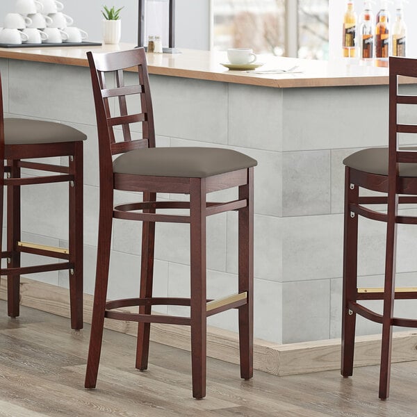Three Lancaster Table & Seating mahogany wood bar stools with taupe vinyl seats at a bar counter.