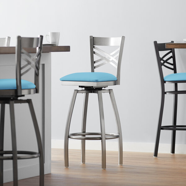 A close-up of a Lancaster Table & Seating clear coat finish cross back swivel bar stool with blue vinyl padded seat.