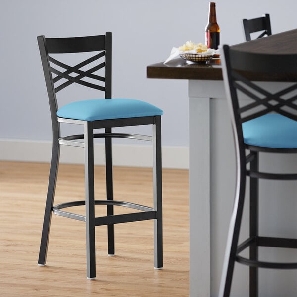 A Lancaster Table & Seating black cross back bar stool with a blue vinyl padded seat next to a table.