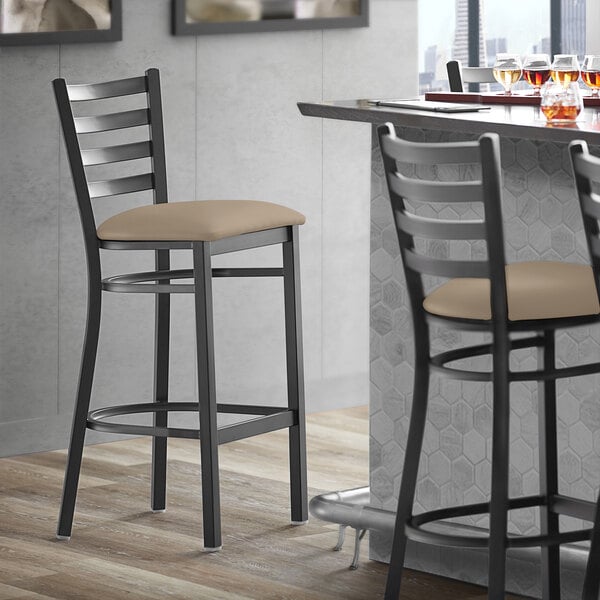 A Lancaster Table & Seating black finish ladder back bar stool with taupe vinyl padded seat next to a table.