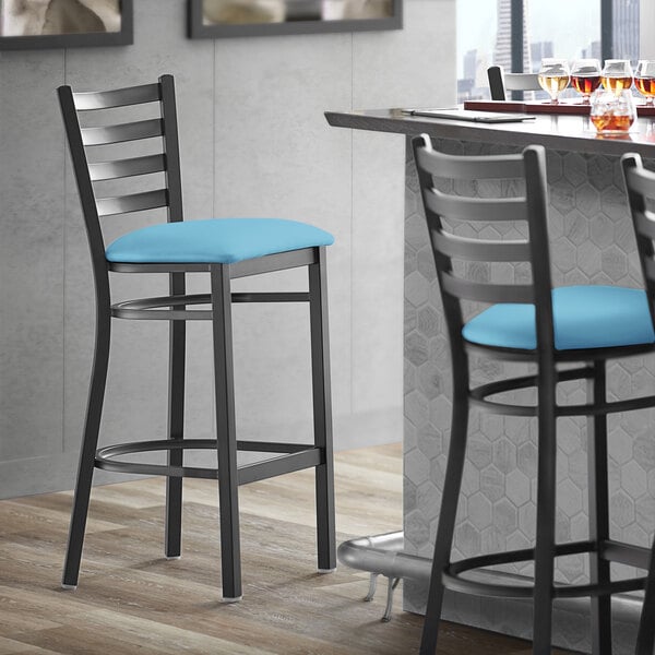 A Lancaster Table & Seating black ladder back bar stool with a blue vinyl padded seat next to a table.