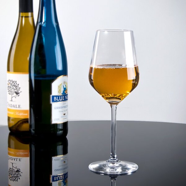 A Stolzle white wine glass filled with yellow liquid next to two bottles of wine.