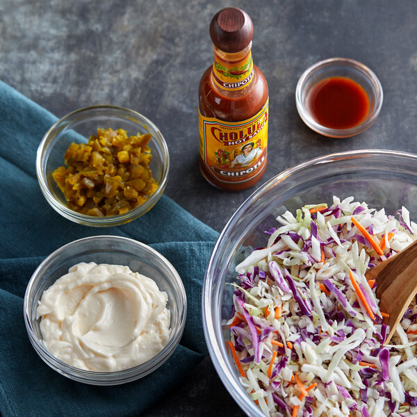 A bowl of shredded cabbage and carrots next to a bowl of red liquid with a bottle of Cholula Chipotle Hot Sauce.