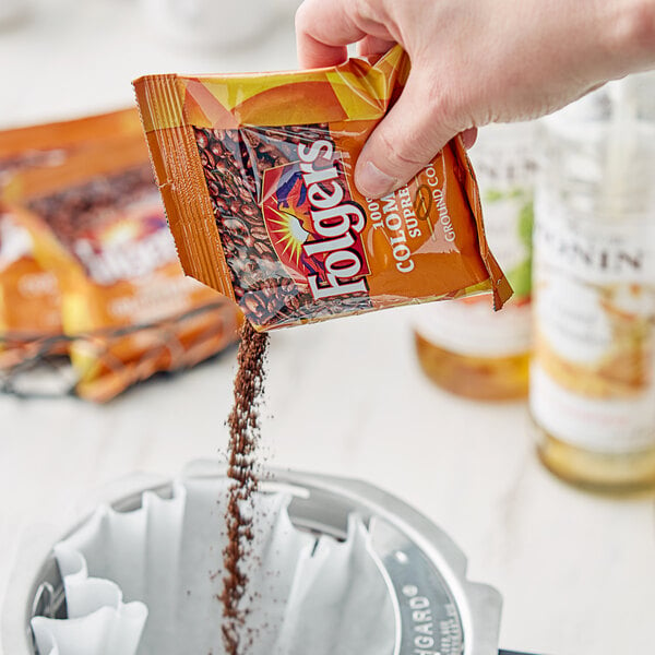 A hand pouring Folgers Colombian Supreme coffee from a packet into a coffee maker.