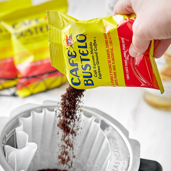 A hand pouring Cafe Bustelo Espresso ground coffee from a yellow and red packet into a coffee filter.