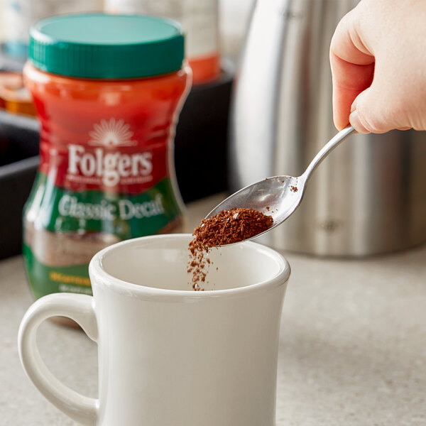 A person pouring Folgers Classic Decaf Instant Coffee into a mug.