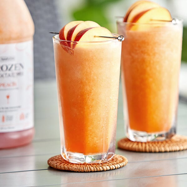 Two glasses of Narvon Peach frozen cocktail mix and orange juice on a table with fruit slices.