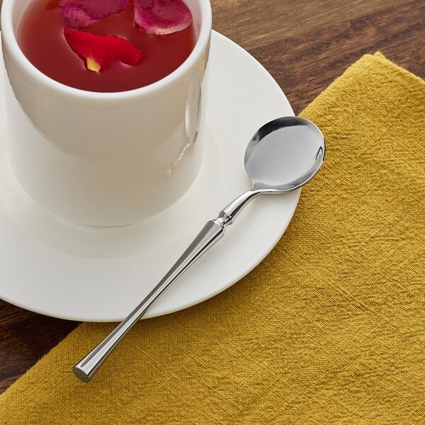 An Acopa stainless steel demitasse spoon on a saucer with a cup of liquid and rose petals.