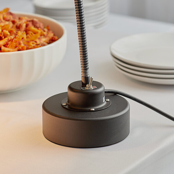 A black Avantco weighted base heat lamp over a bowl of pasta on a table.
