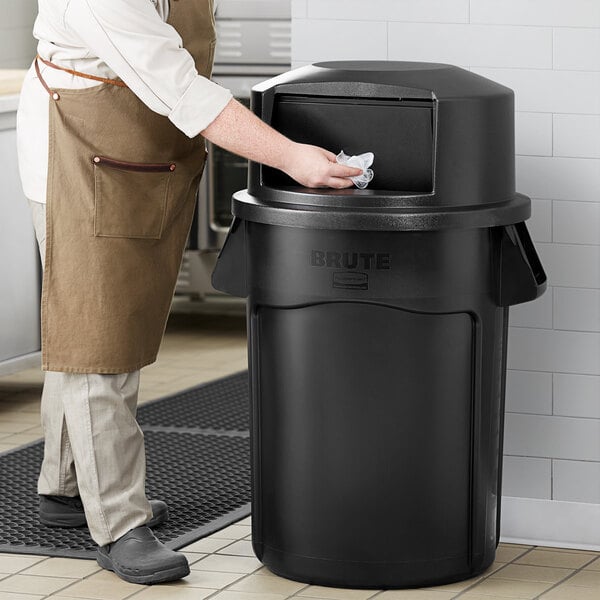 A person in a black apron and gloves cleaning a Rubbermaid Black BRUTE trash can.