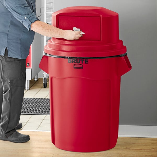 A man standing next to a red Rubbermaid BRUTE trash can with a red dome top.