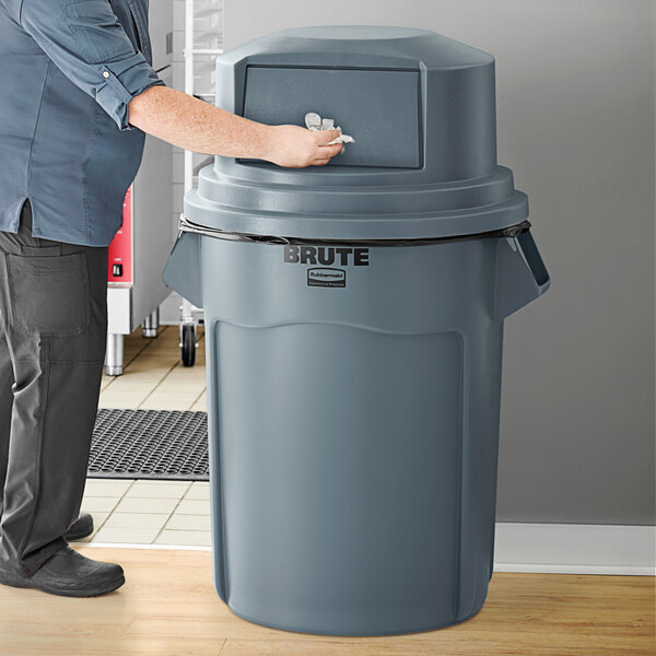 A person opening a Rubbermaid BRUTE 55 gallon grey trash can with a grey round dome top.