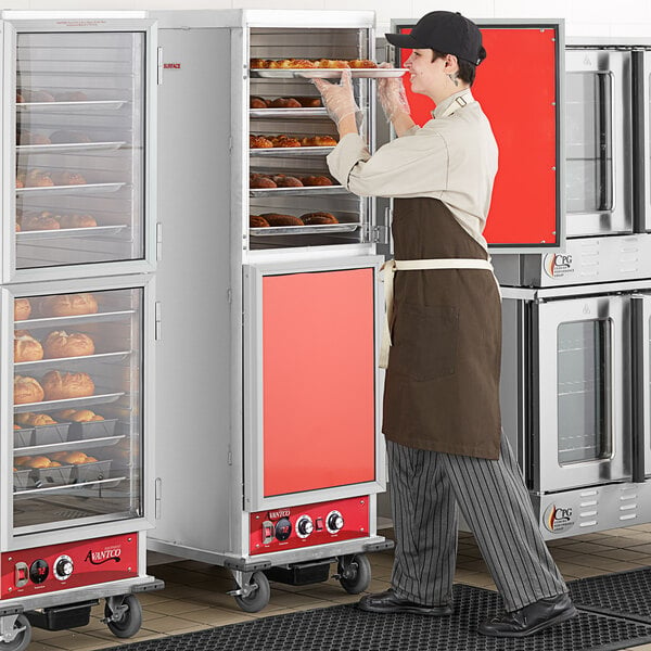 A man in a brown apron standing in front of a large stainless steel holding and proofing cabinet with solid doors.