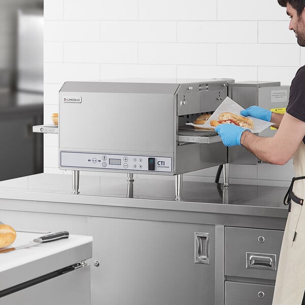 A man in a white apron using a Lincoln countertop conveyor oven to prepare a sandwich.